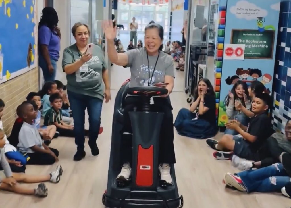 Ms. Ha, riding floor cleaner down hallway with students cheering