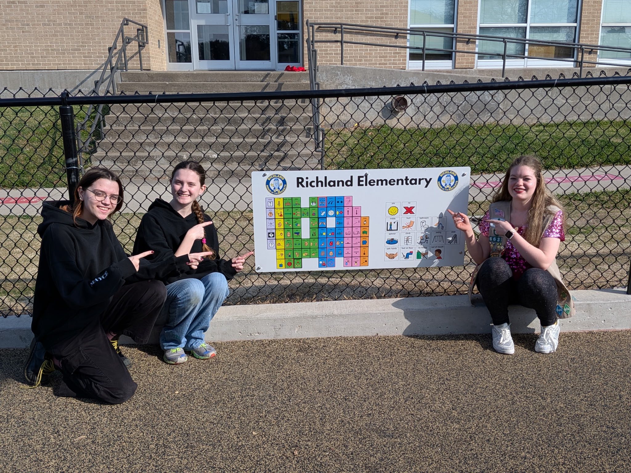 Three students pointing to visual communication board at Richland Elementary hung on a playground fence