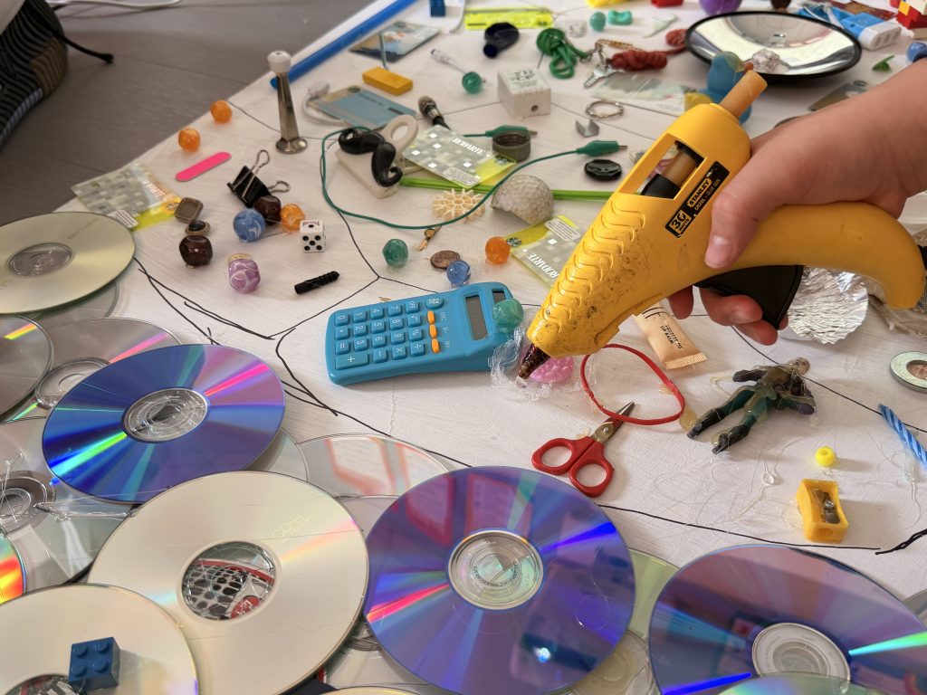 found objects being glued to board including calculator, chapstick, cds, bouncy balls, keys and more