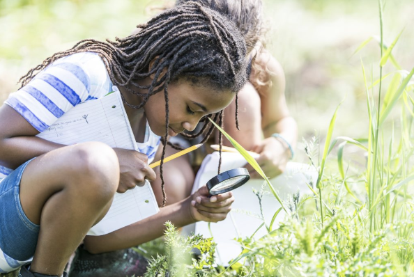children outdoors, one is holding a magnifying glass looking at grass while holding a pencil and paper