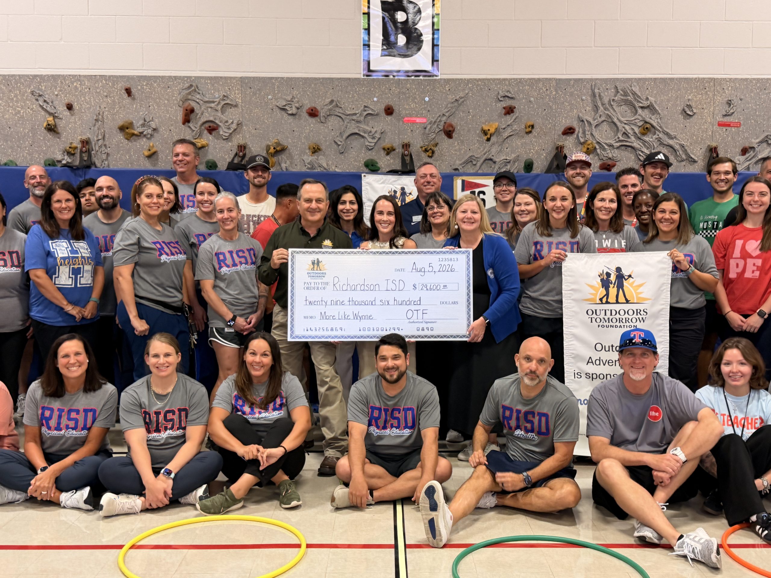 A group of people, many wearing "RISD Physical Education" shirts, pose for a photo in a gymnasium with a climbing wall in the background and big check.