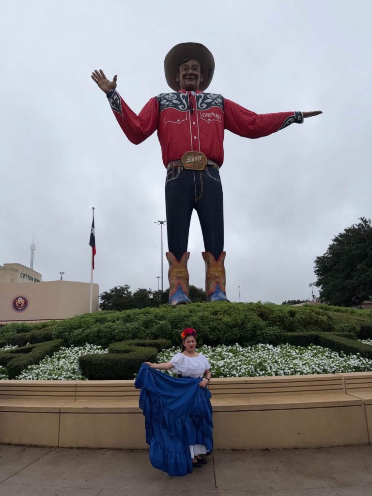 Brianna Mora in traditional Folklorico dress posing in front of Big Tex at the State Fair